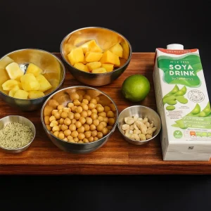 Smoothie ingredients arranged on a wooden board: two metal bowls containing diced pineapple, a bowl of chickpeas, a small bowl of cashews, a small bowl of hemp seeds, a whole lime, and a carton of Sainsbury's Unsweetened Soya Drink.