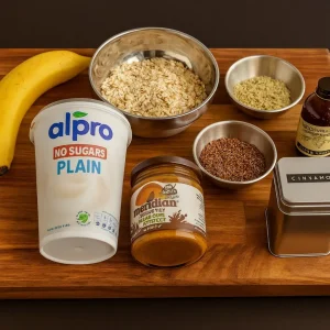 Ingredients laid out on a wooden cutting board: a yellow banana, a container of Alpro No Sugars Plain yogurt, a bowl of rolled oats, small bowls of hemp seeds and flax seeds, a jar of nut butter, and a small dark glass bottle of extract, next to a silver tin labeled "CINNAMON."