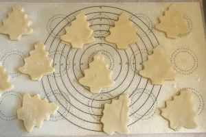 Unbaked sugar cookie dough cut into Christmas tree shapes and arranged on a silicone baking mat.

