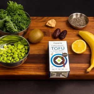Various ingredients for a green smoothie displayed on a wooden board, including a large bowl of curly kale, a bowl of shelled edamame, a kiwi, two dates, a piece of ginger, a box of Clearspring Organic Tofu, a small bowl of chia seeds, half a lemon, and a banana.