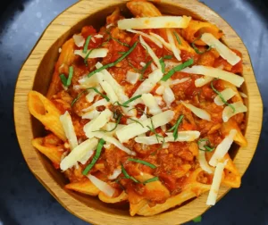 A wooden bowl of penne pasta tossed in a rich red bolognese sauce, topped with shredded white cheese and fresh basil or oregano.