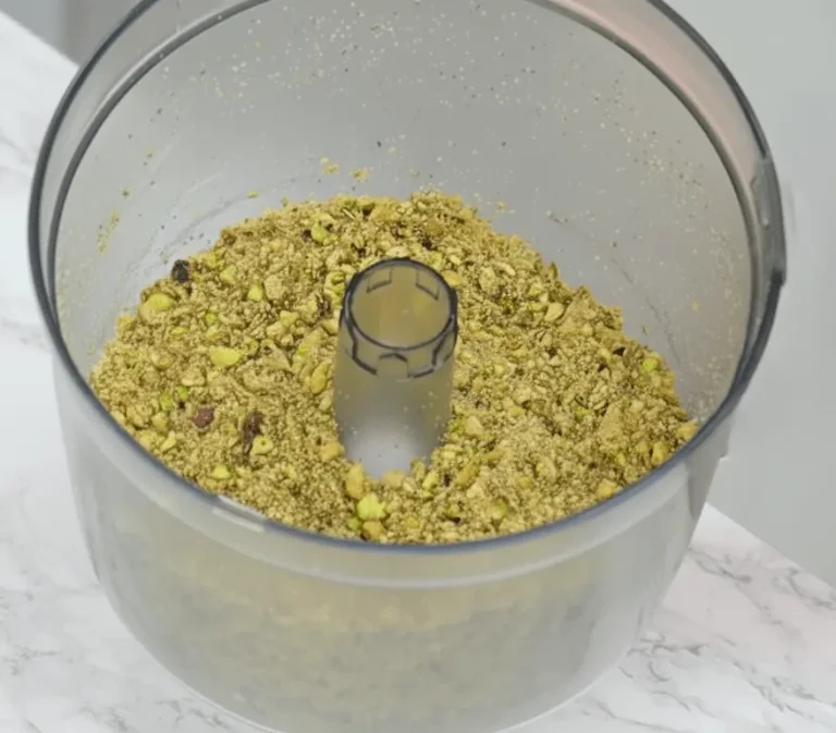 A close-up shot of a food processor bowl filled with coarsely chopped pistachios and other nuts, sitting on a white marble countertop.