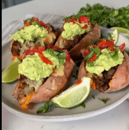 Several baked sweet potatoes split open and stuffed with seasoned meat and melted cheese, topped with guacamole, salsa or hot sauce, and cilantro, served with lime wedges.
