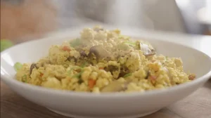 A white bowl filled with steaming tofu scramble, mixed with mushrooms and green onions, suggestive of a vegan breakfast dish.