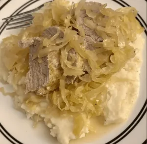 A white plate with a serving of mashed potatoes topped with slices of meat and a generous portion of cooked cabbage or sauerkraut.