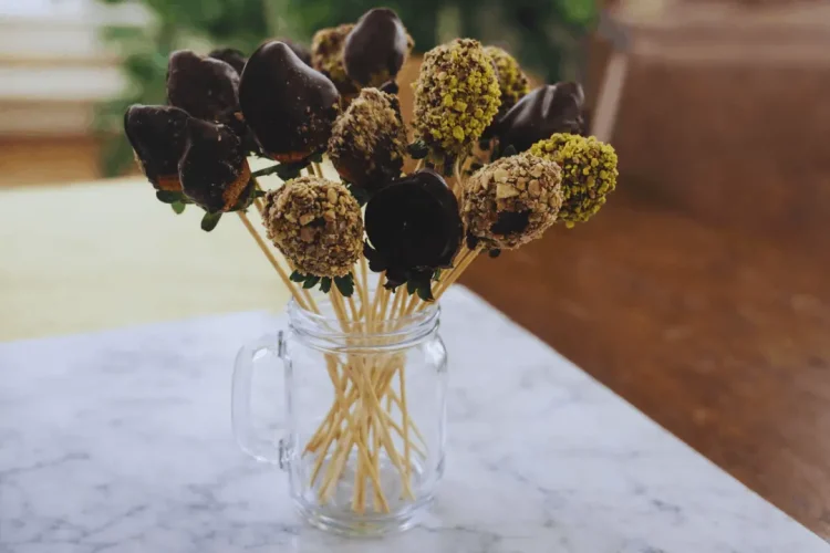 A close-up of a completed edible fruit bouquet. Several dark chocolate-covered strawberries, some plain and some coated in crushed green nuts, are on long wooden skewers and arranged in a clear glass mason jar. The bouquet sits on a white marble surface.