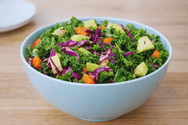 A large light-blue bowl filled with a fresh salad made of chopped curly kale, shredded red cabbage, diced carrots, and chunks of green apple. The bowl is sitting on a light-colored wooden surface.