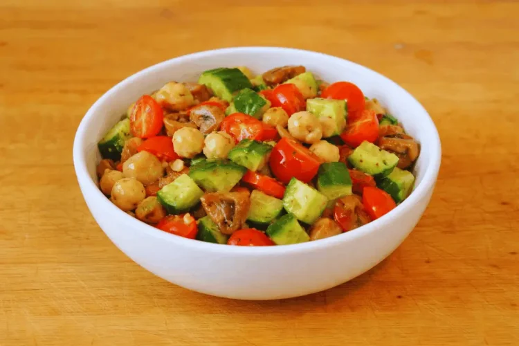 A white bowl filled with a vibrant chickpea salad containing halved cherry tomatoes, diced cucumbers, and sliced mushrooms. The bowl sits on a wooden cutting board.