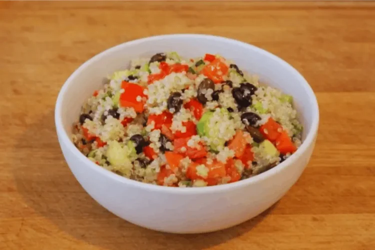 A white bowl containing a quinoa salad mixed with black beans, diced red tomatoes, and cucumbers, presented on a wooden table.