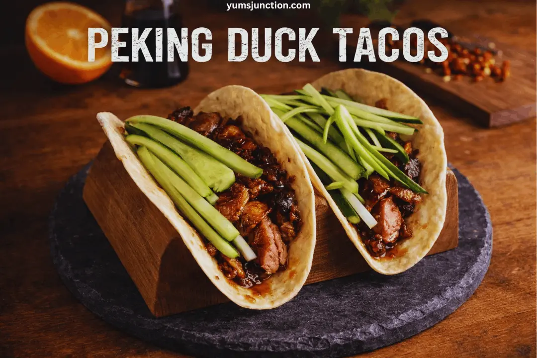 A professional food photograph of two Peking duck tacos in soft tortillas, filled with glossy, dark-glazed duck meat and topped with fresh julienned cucumbers and spring onions. The tacos are held upright in a wooden taco rack sitting on a dark slate platter. Large, bold white text across the top reads "PEKING DUCK TACOS" with the website "yumsjunction.com" above it.