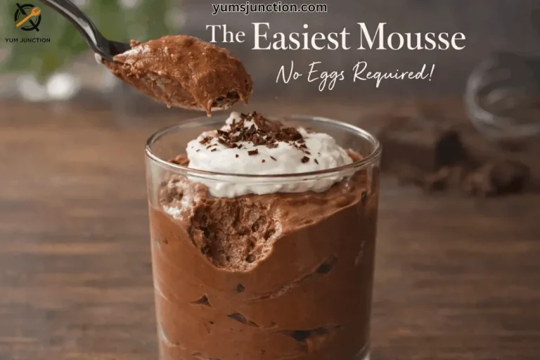 A landscape-oriented promotional image. In the center, a clear glass is filled with a thick, aerated chocolate mousse topped with a large dollop of white whipped cream and a sprinkle of dark chocolate shavings. A dark metal spoon is lifting a generous, textured scoop of mousse from the glass. The title "The Easiest Mousse" appears in white serif font, with "No Eggs Required!" in a white script below it. The background is a blurred dark wooden surface. The Yum Junction logo is in the top left, and the URL yumsjunction.com is at the top center.