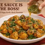 A horizontal food graphic with the headline "THE SAUCE IS THE BOSS!" in bold dark brown text, followed by "Ultra-Crispy Garlic Herb Potatoes." In the center, a hand is captured pouring a vibrant green herb-flecked oil from a small glass jar onto a large white bowl of crispy roasted potatoes. The potatoes are garnished with parmesan and parsley and sit on a red cloth napkin. The Yum Junction logo is in the top left corner, and the website URL "yumsjunction.com" is centered at the top.