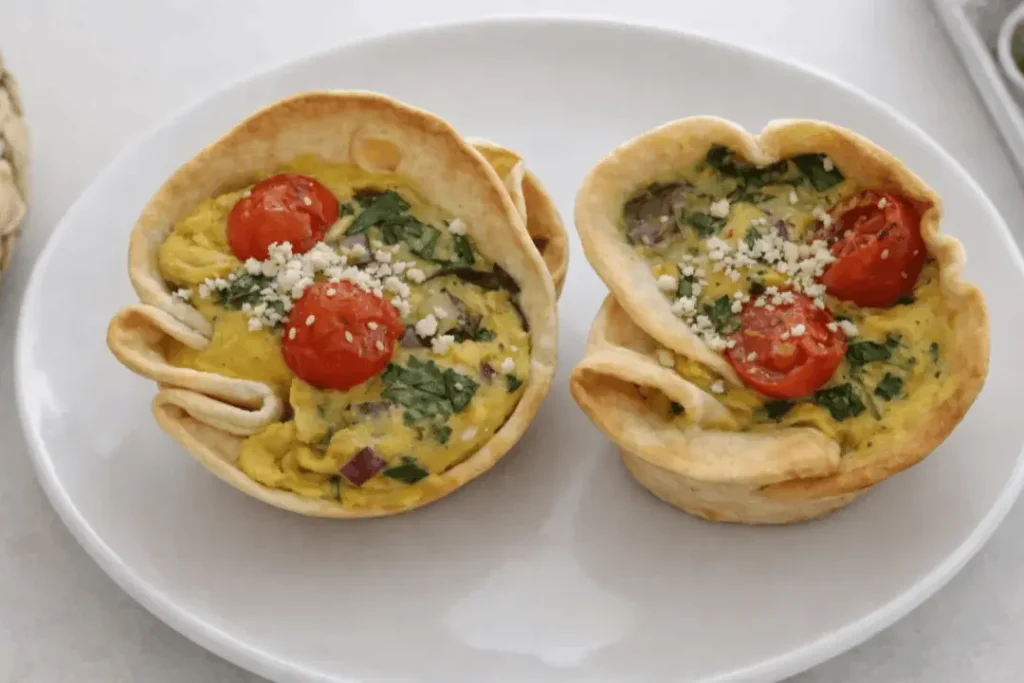 Two golden mini egg cups filled with scrambled eggs, cherry tomatoes, red onion, and feta cheese, served on a white plate