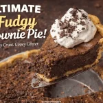 A slice of ultimate fudgy brownie pie with crispy crust and gooey center being lifted on a pie server, topped with whipped cream and chocolate shavings