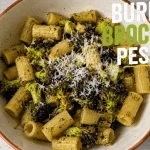 A top-down view of a bowl filled with rigatoni pasta tossed in vibrant green pesto, topped with charred burnt broccoli florets and freshly grated parmesan cheese, labeled with the text "Burnt Broccoli Pesto"
